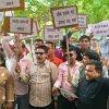 Group of people at a protest holding placards with Hindi messages, shouting together outdoors under trees.