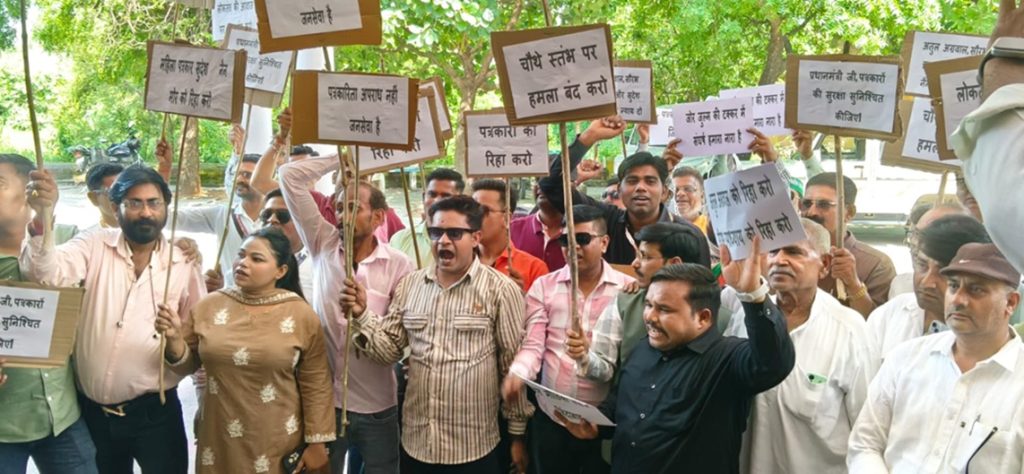 Group of people at a protest holding placards with Hindi messages, shouting together outdoors under trees.
