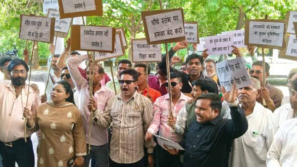 Group of people at a protest holding placards with Hindi messages, shouting together outdoors under trees.