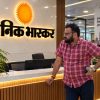 Bearded man in a red plaid shirt stands at a marble reception desk with a sun logo in Hindi signage behind him.