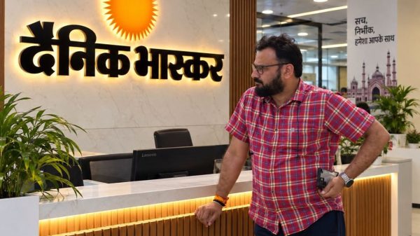 Bearded man in a red plaid shirt stands at a marble reception desk with a sun logo in Hindi signage behind him.