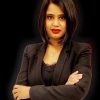 Professional woman with dark hair in a black blazer, arms crossed, looking at the camera against a dark background.