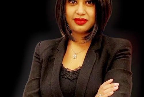 Professional woman with dark hair in a black blazer, arms crossed, looking at the camera against a dark background.