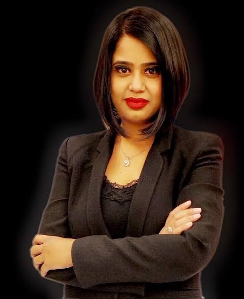 Professional woman with dark hair in a black blazer, arms crossed, looking at the camera against a dark background.