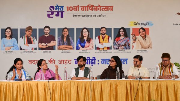 Panelists seated behind a long table with microphones and water bottles, in front of a large event banner featuring portraits and Hindi text.