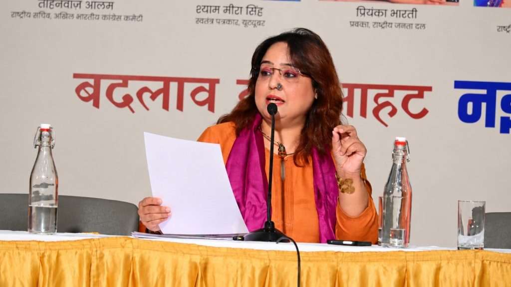 mera rang_3 | No. 1 Indian Media News Portal Panel speaker: woman in orange kurta and pink scarf reads from papers into a microphone at a conference table with water bottles nearby