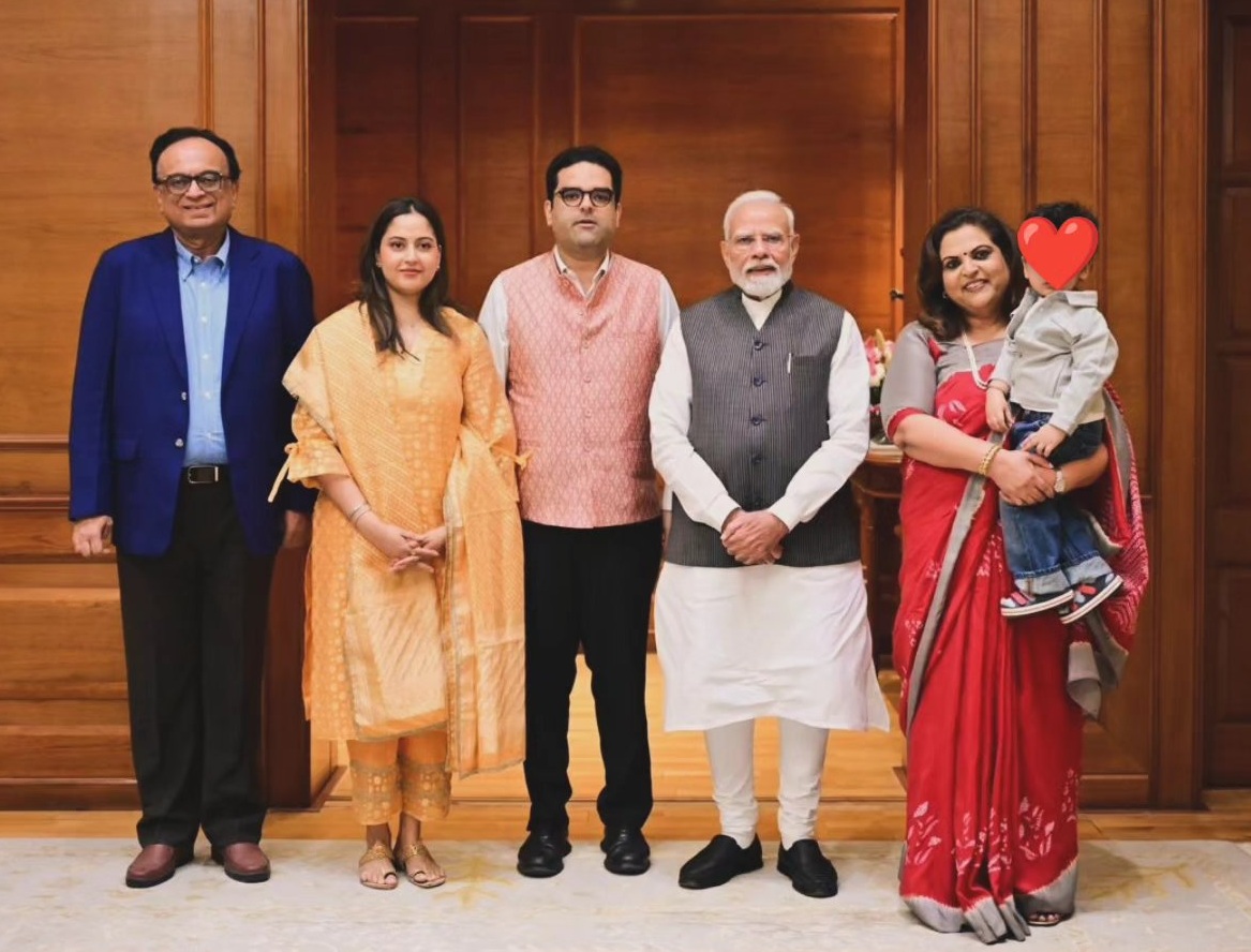 Group of five adults posing for a formal photo in a wooden-paneled room, one woman holding a child with a heart sticker over the child's face.