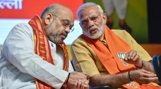 Two men seated on a stage in traditional attire, both wearing orange scarves, leaning toward each other as they talk during a public event.