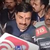 Man with a mustache in a dark suit speaks to reporters as microphones from various news outlets loom around him in a crowded press scrum.