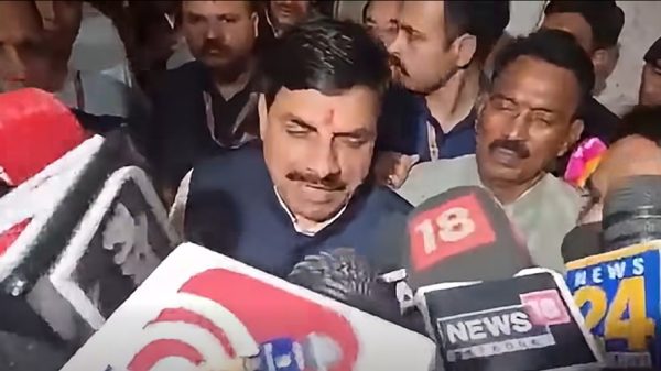 Man with a mustache in a dark suit speaks to reporters as microphones from various news outlets loom around him in a crowded press scrum.