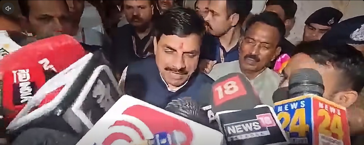 Man with a mustache in a dark suit speaks to reporters as microphones from various news outlets loom around him in a crowded press scrum.