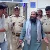 Bearded man in teal kurta and light scarf speaks while being escorted by two uniformed police officers in a hallway.