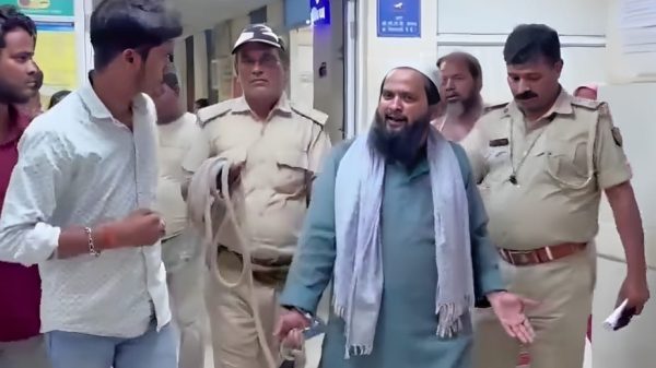 Bearded man in teal kurta and light scarf speaks while being escorted by two uniformed police officers in a hallway.