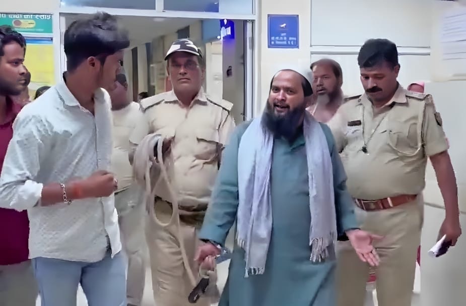 Bearded man in teal kurta and light scarf speaks while being escorted by two uniformed police officers in a hallway.