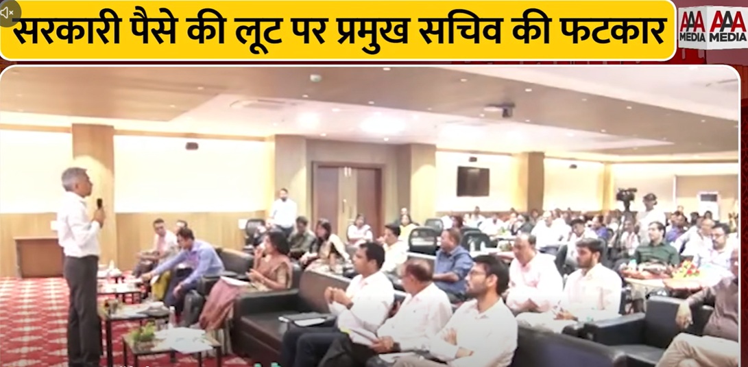 Speaker addressing a conference in a hall as attendees listen; a bold Hindi banner about government funds misuse is visible at the top.