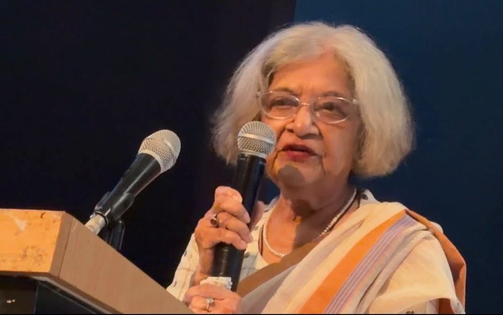 mradula | No. 1 Indian Media News Portal Elderly woman with short white hair in a cream sari speaks into two microphones at a podium.