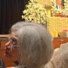 Elderly woman with gray hair and glasses seated at a memorial service, with white flower arrangements and a portrait in the background.