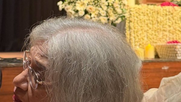 Elderly woman with gray hair and glasses seated at a memorial service, with white flower arrangements and a portrait in the background.
