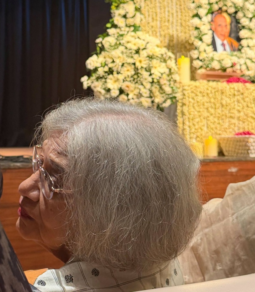 Elderly woman with gray hair and glasses seated at a memorial service, with white flower arrangements and a portrait in the background.