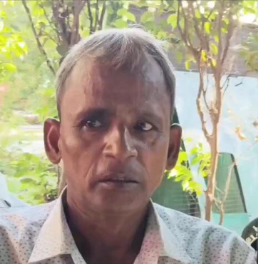 Portrait of a man with short hair wearing a light shirt, seated outdoors with trees in the background.