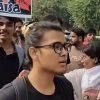 Woman with glasses and black shirt walking in a protest crowd, foreground focus with a colorful protest sign in the background. Moment captured outdoors among other protesters and trees.