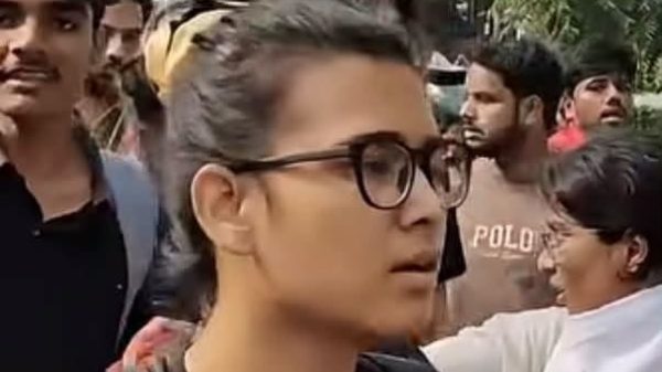 Woman with glasses and black shirt walking in a protest crowd, foreground focus with a colorful protest sign in the background. Moment captured outdoors among other protesters and trees.