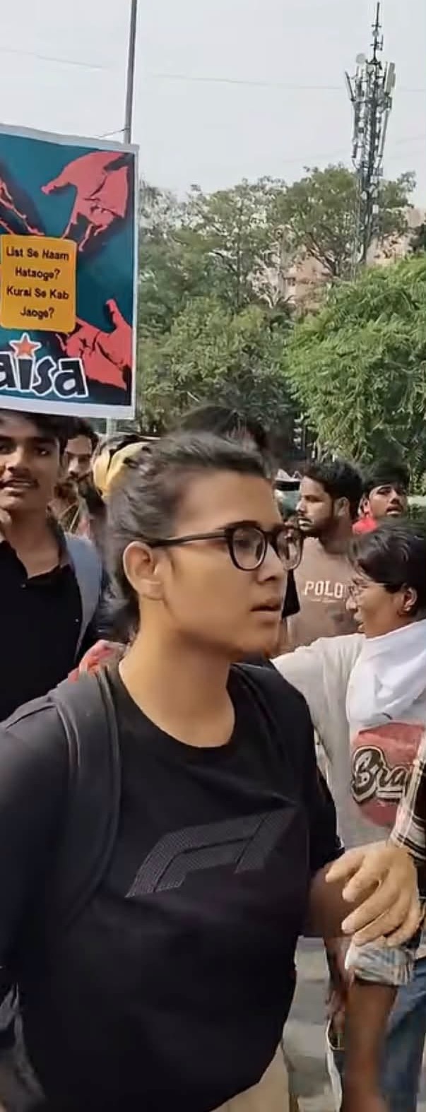 Woman with glasses and black shirt walking in a protest crowd, foreground focus with a colorful protest sign in the background. Moment captured outdoors among other protesters and trees.