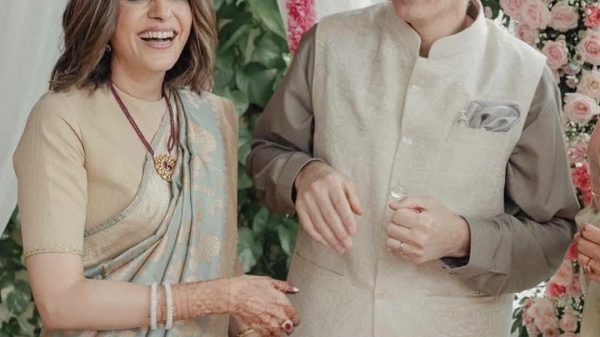 Joyful Indian couple at a wedding, woman in pale blue saree with mehndi, man in beige kurta, pink floral backdrop and petals framing them.