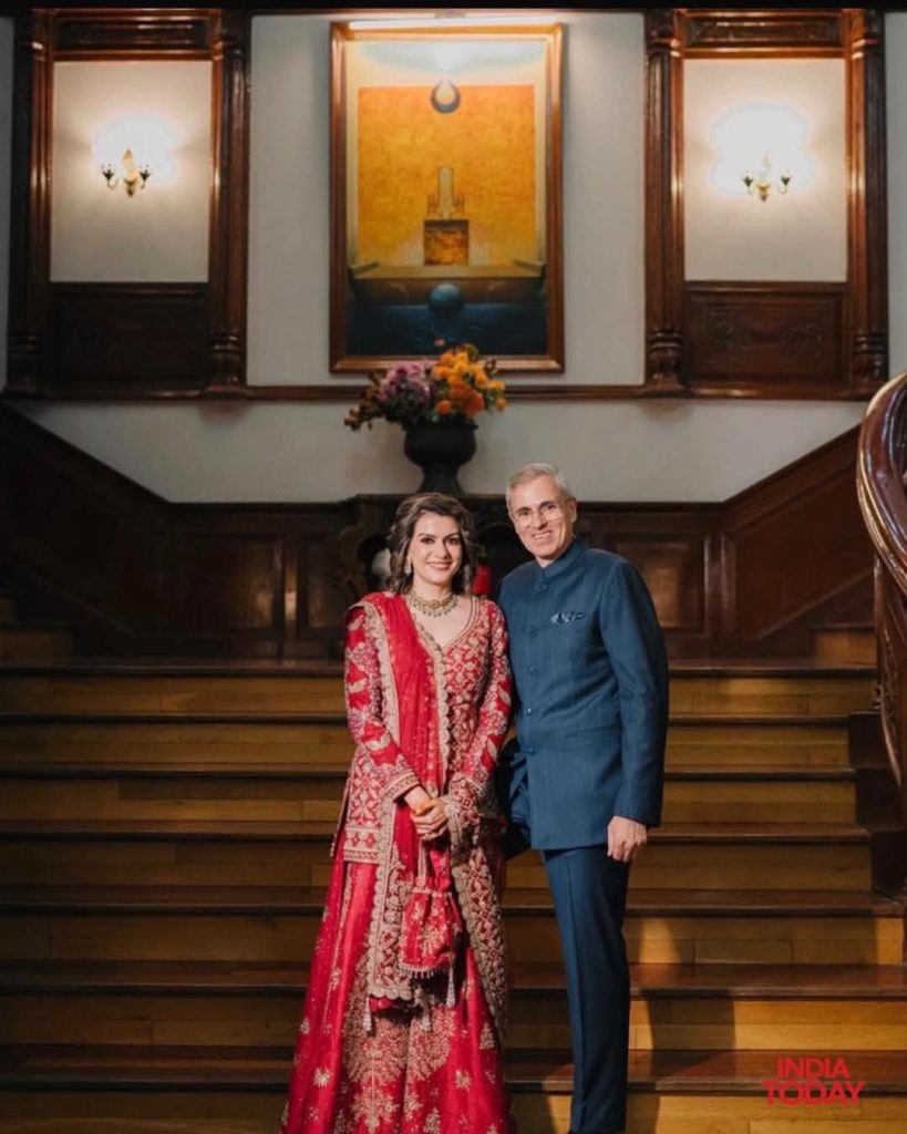 nidhi_omr-1 | No. 1 Indian Media News Portal Couple posing on a grand wooden staircase indoors; woman wearing a red embroidered traditional dress and jewelry, beside a man in a blue suit.