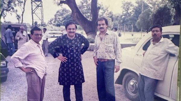Four men stand together outdoors beside a white vintage car, with trees in the background.