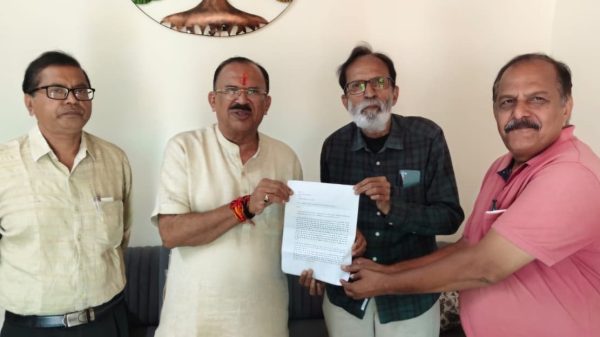 Four Indian men stand indoors, with one in a cream kurta presenting a document to a man in a green plaid shirt while another man in a pink polo stands beside them.