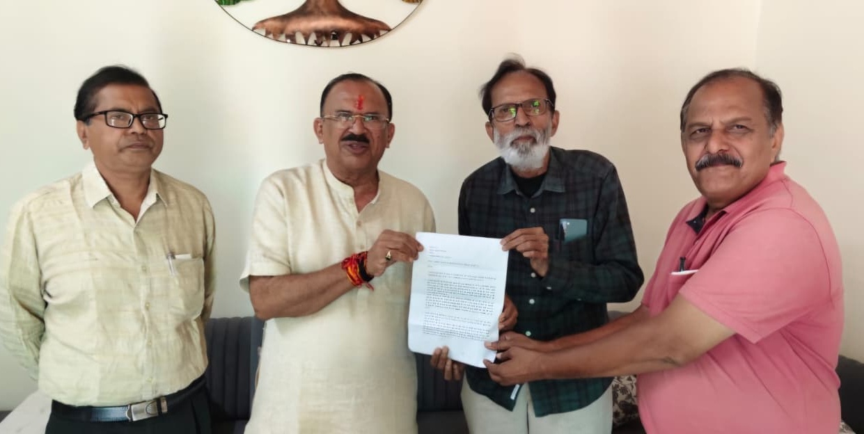 Four Indian men stand indoors, with one in a cream kurta presenting a document to a man in a green plaid shirt while another man in a pink polo stands beside them.