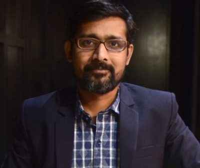 Man with glasses and beard wearing a navy blazer and checkered shirt, looking at the camera in a dim background environment