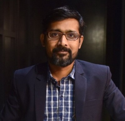 pavan deshpande | No. 1 Indian Media News Portal Man with glasses and beard wearing a navy blazer and checkered shirt, looking at the camera in a dim background environment
