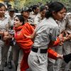 Policewomen in khaki uniforms restrain a woman in an orange sari as a reporter holds a microphone nearby in a street scene.