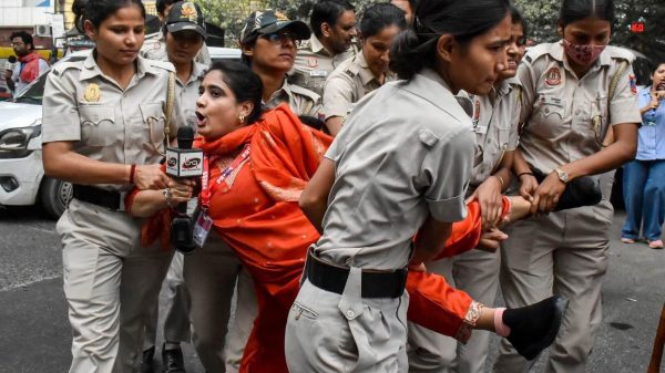 Policewomen in khaki uniforms restrain a woman in an orange sari as a reporter holds a microphone nearby in a street scene.