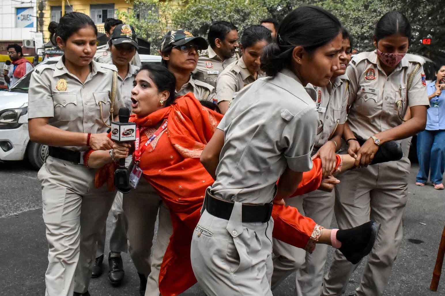pragya | No. 1 Indian Media News Portal Policewomen in khaki uniforms restrain a woman in an orange sari as a reporter holds a microphone nearby in a street scene.