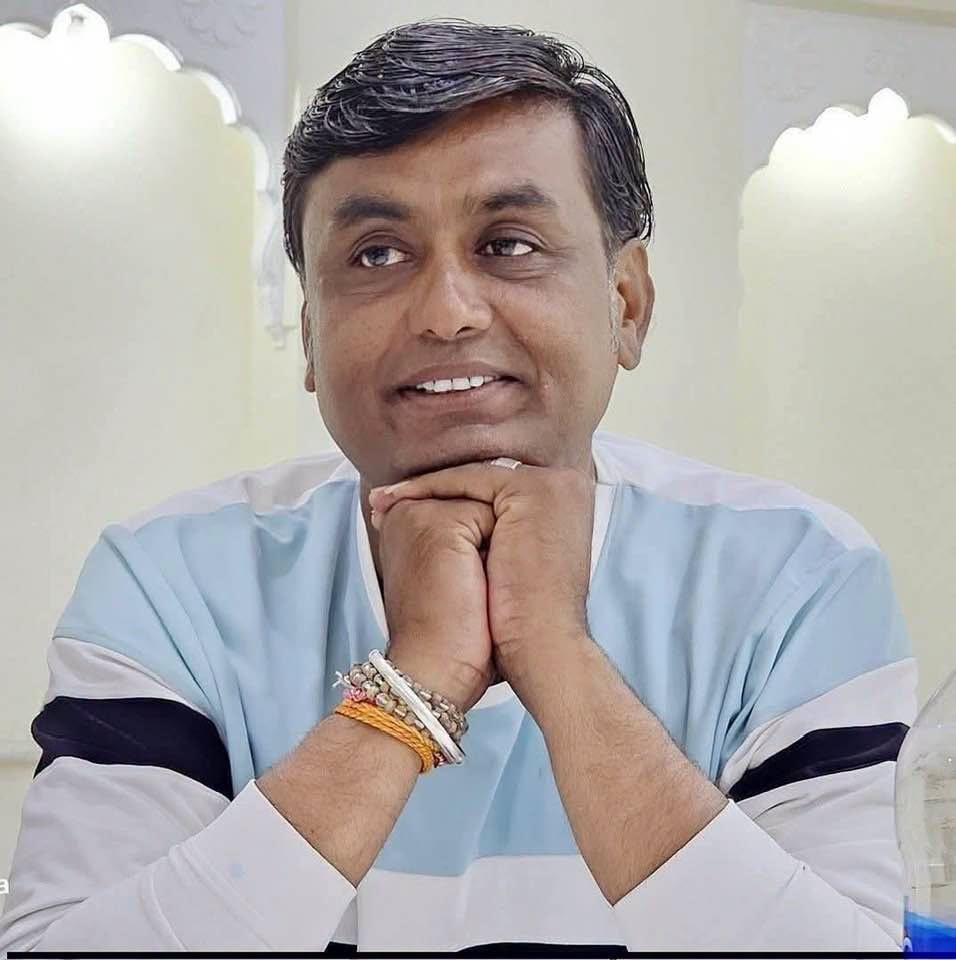 praveen | No. 1 Indian Media News Portal Man in a light blue and white striped shirt rests his chin on clasped hands, smiling softly indoors, wearing bracelets on one wrist.