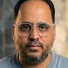 Close-up of a man with short dark hair and beard wearing clear safety glasses, looking at the camera against a blurred background.