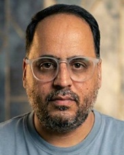 Close-up of a man with short dark hair and beard wearing clear safety glasses, looking at the camera against a blurred background.