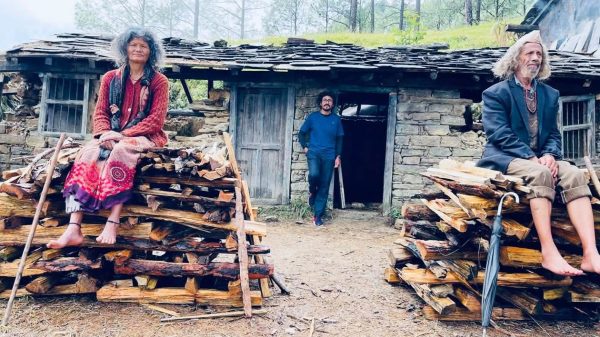 Three adults sit on stacked firewood outside a rustic stone hut in a wooded area; a woman in red on the left, a man in blue near the doorway, another man in a blazer on the right.