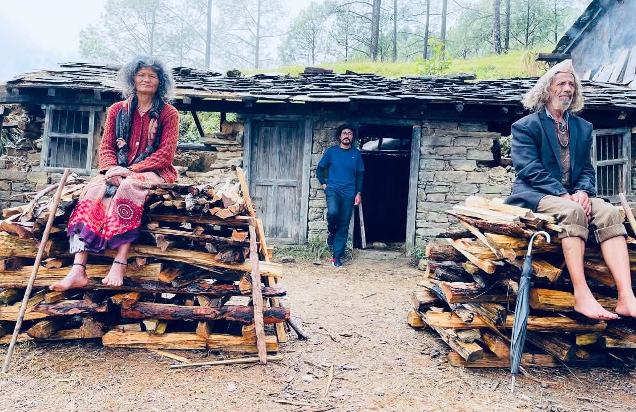 Three adults sit on stacked firewood outside a rustic stone hut in a wooded area; a woman in red on the left, a man in blue near the doorway, another man in a blazer on the right.
