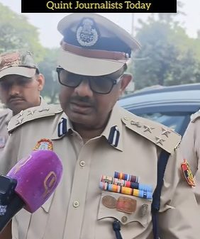 Police officer in tan uniform and cap speaks to reporters, sunglasses on, microphone held near him; text banner overhead reads: 'How Delhi Police Detained, Heckled, Tailled, And Questioned Quint Journalists Today'