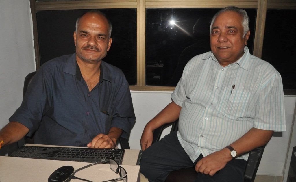 raju_buddhi | No. 1 Indian Media News Portal Two middle-aged men sit at a desk with a laptop, smiling in an indoor office at night.