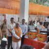 An Indian official in an orange vest stands at a table with two orange donation/ceremony machines while a crowd holds up certificates and takes photos at an event stall.