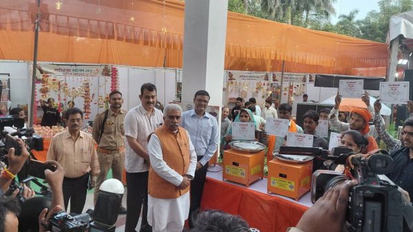 An Indian official in an orange vest stands at a table with two orange donation/ceremony machines while a crowd holds up certificates and takes photos at an event stall.