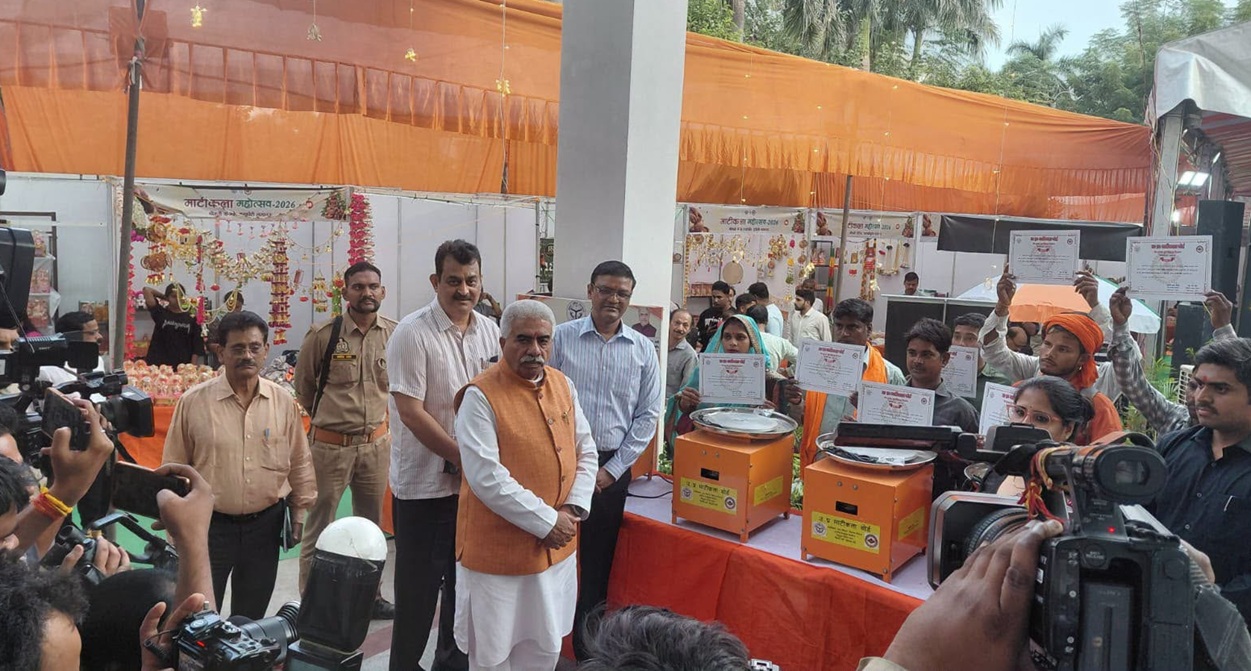 rakesh | No. 1 Indian Media News Portal An Indian official in an orange vest stands at a table with two orange donation/ceremony machines while a crowd holds up certificates and takes photos at an event stall.