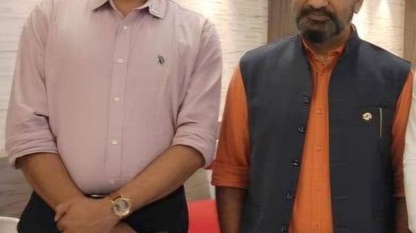 Two men stand indoors side by side, posing for a photo: one in a light pink button-down shirt and black trousers, the other in an orange shirt with a dark vest and khaki pants.