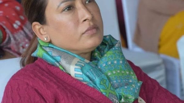 South Asian woman at an event, wearing a pink cardigan and green patterned scarf, with a red tilak on her forehead, looking to the side.