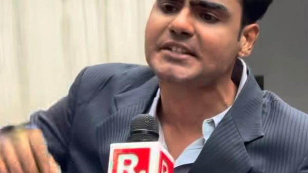 Male news reporter in a pinstripe suit speaking into a mic with a red R logo.